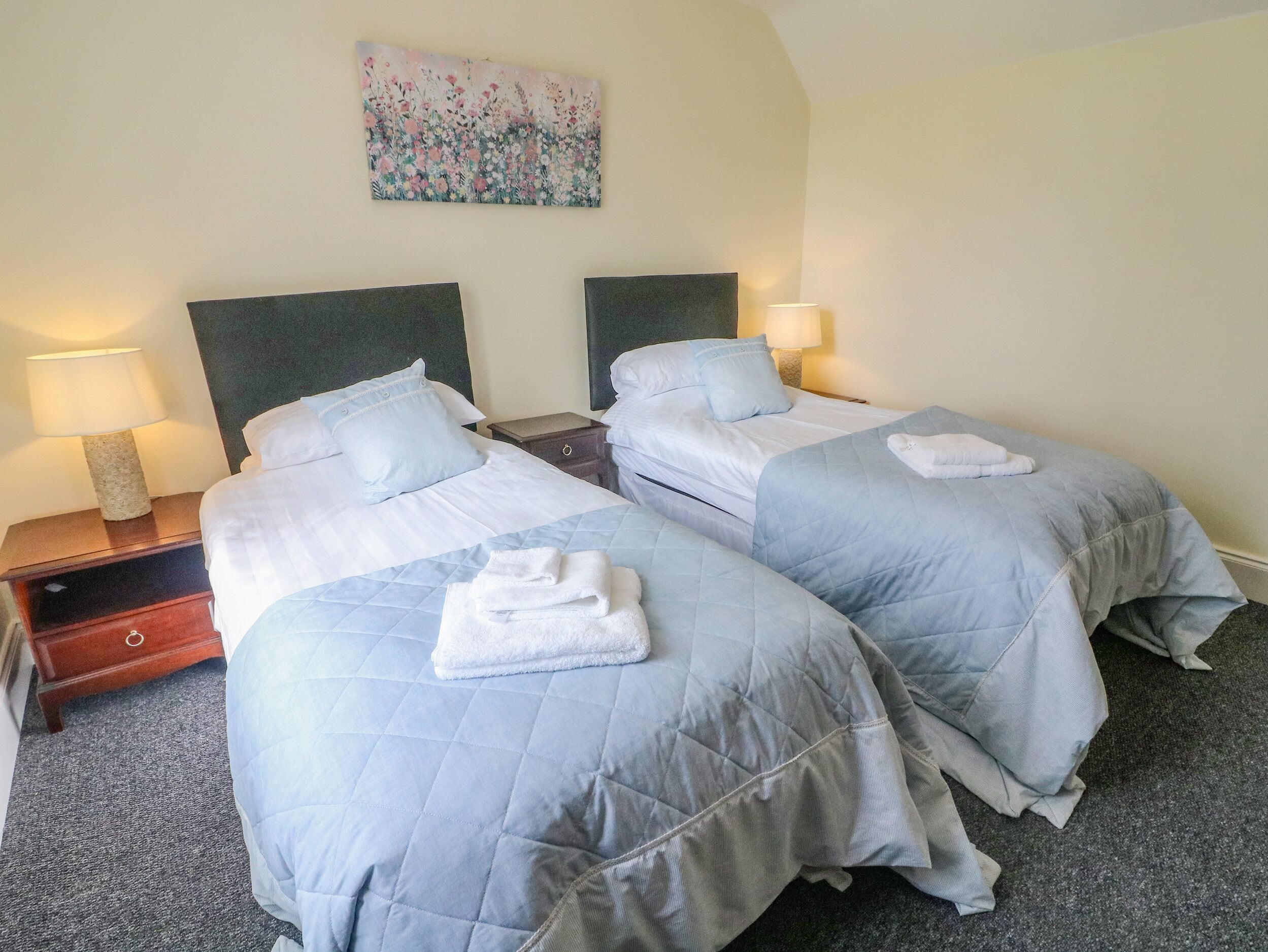 A hotel room with two beds, each with a blue quilted bedspread and white pillows. There are bedside tables with lamps, a wooden nightstand, and a painting on the wall.
