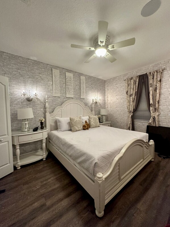 A bedroom with a white bed, a nightstand with a lamp, a ceiling fan, and a brick-patterned wall.
