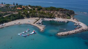 Luftaufnahme einer Küstenregion mit einem Yachthafen, klarem, blauem Wasser und einem kleinen Strand mit Sonnenschirmen.