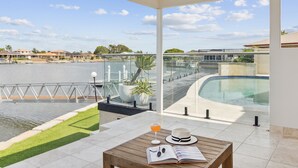 Un balcon donnant sur une piscine, avec une balustrade en verre et une table sur laquelle se trouvent un chapeau, des lunettes de soleil et un verre de jus d’orange.