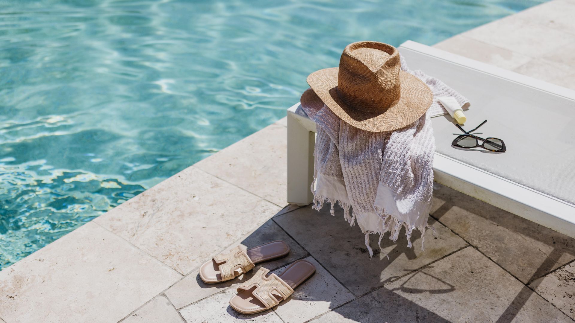 A poolside area with a hat, sunglasses, and sandals on a bench.