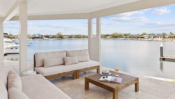 Un espace extérieur aménagé, abrité et doté d’un canapé et d’une table basse, offrant une vue sur un port de plaisance avec ses bateaux et ses maisons.