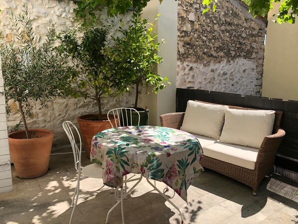 Un espace extérieur aménagé avec un canapé en osier et une table recouverte d’une nappe à motifs floraux. On y trouve également des plantes en pot et un mur de pierre en arrière-plan.