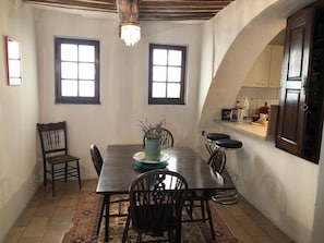 Une salle à manger avec une longue table, des chaises et un lustre. Il y a deux fenêtres et une porte qui donne sur la cuisine.