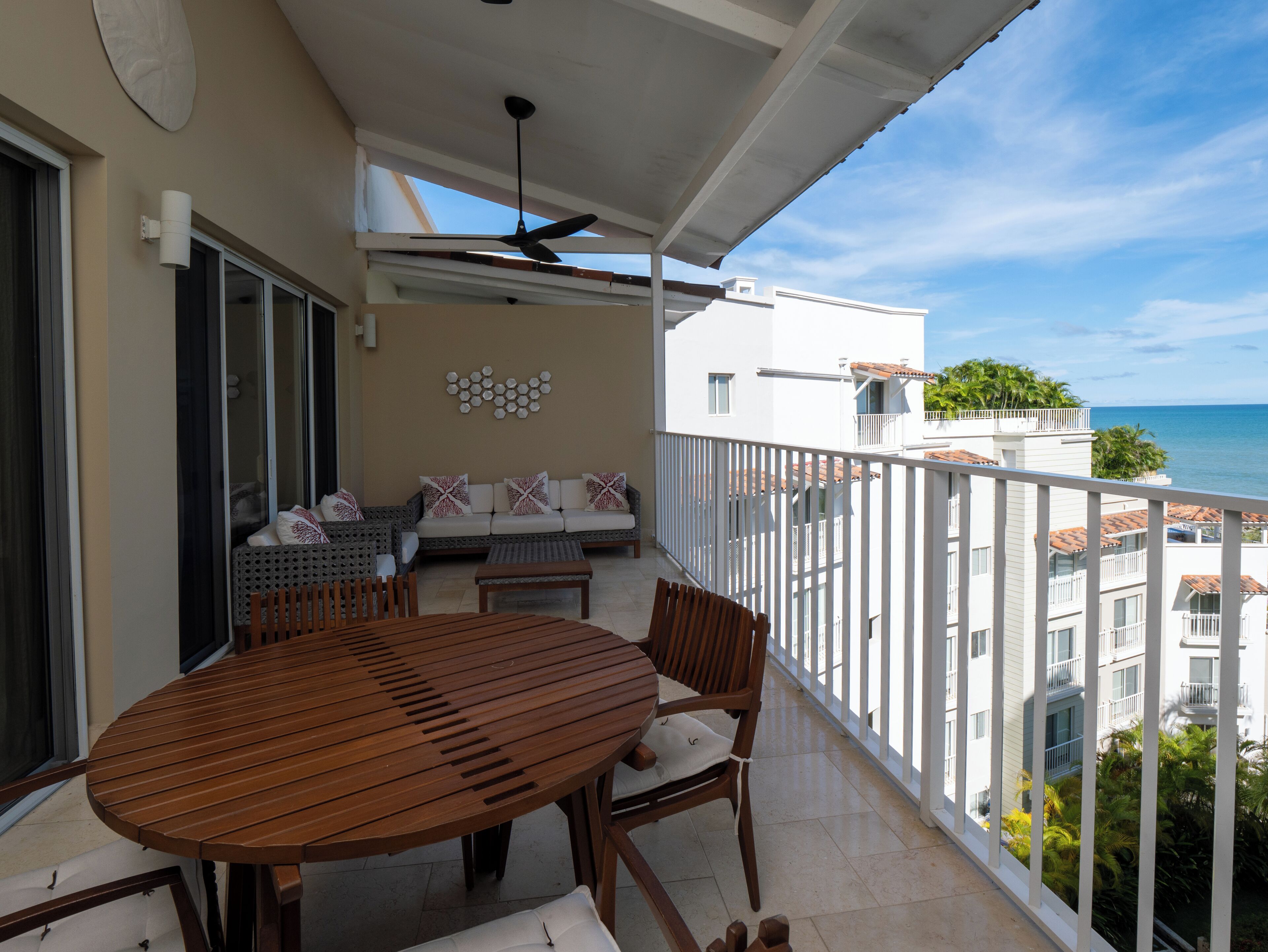 A balcony with a wooden table and chairs, a sofa with patterned cushions, and a ceiling fan.