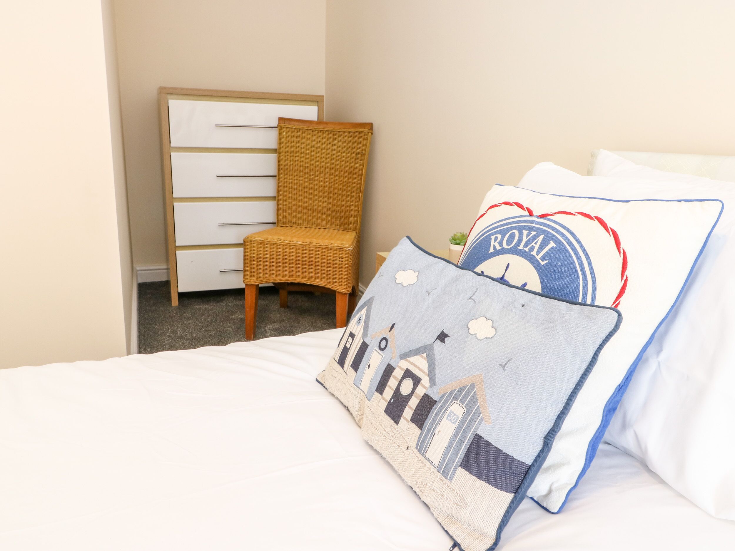A hotel room with a bed, a white dresser, a wicker chair, and decorative pillows.