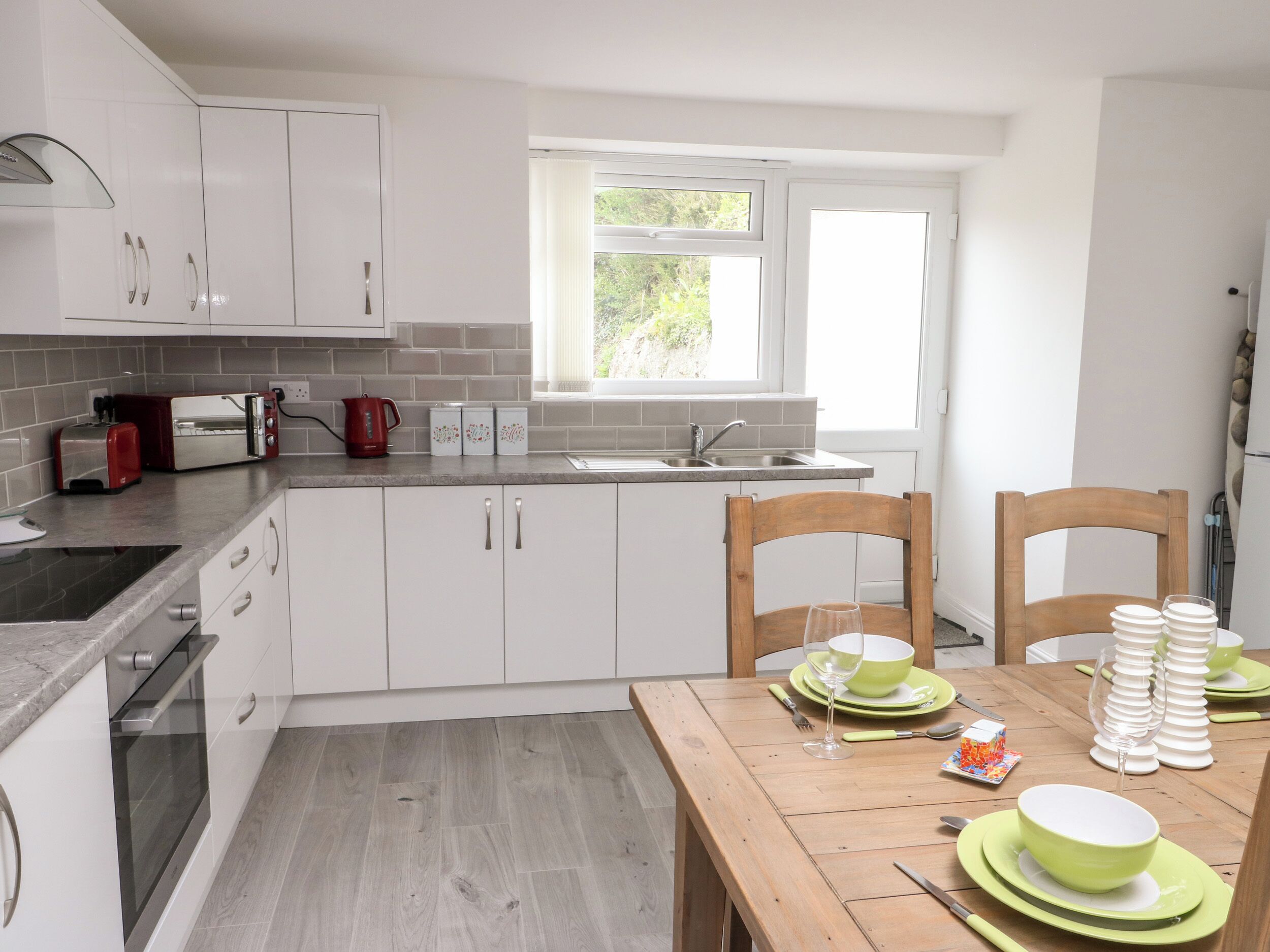 A modern kitchen with white cabinets, a wooden dining table set with green plates, and a window providing natural light.