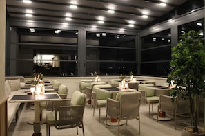 An outdoor dining area with multiple tables and chairs, large windows, and a potted plant.