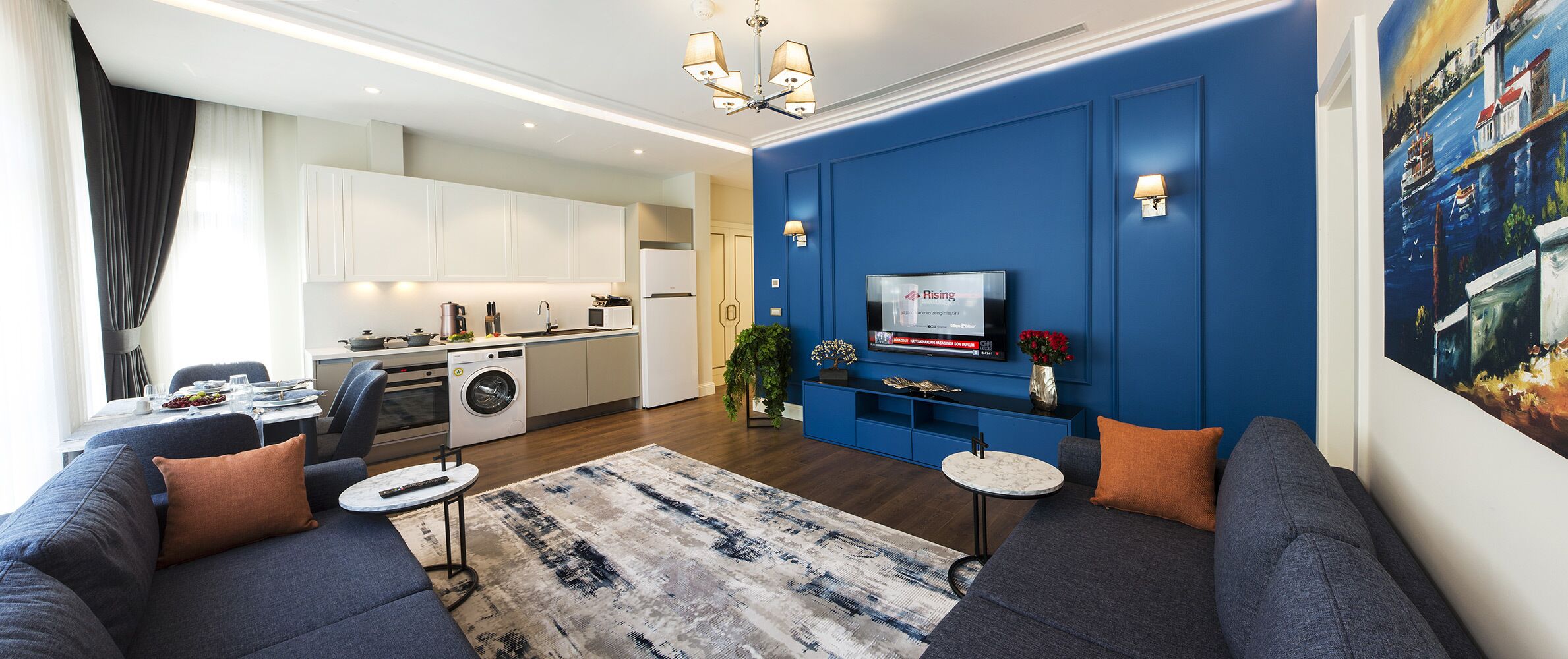 A modern living room with a sofa, a coffee table, and a TV. There is a kitchen in the background with white cabinets and a washing machine.