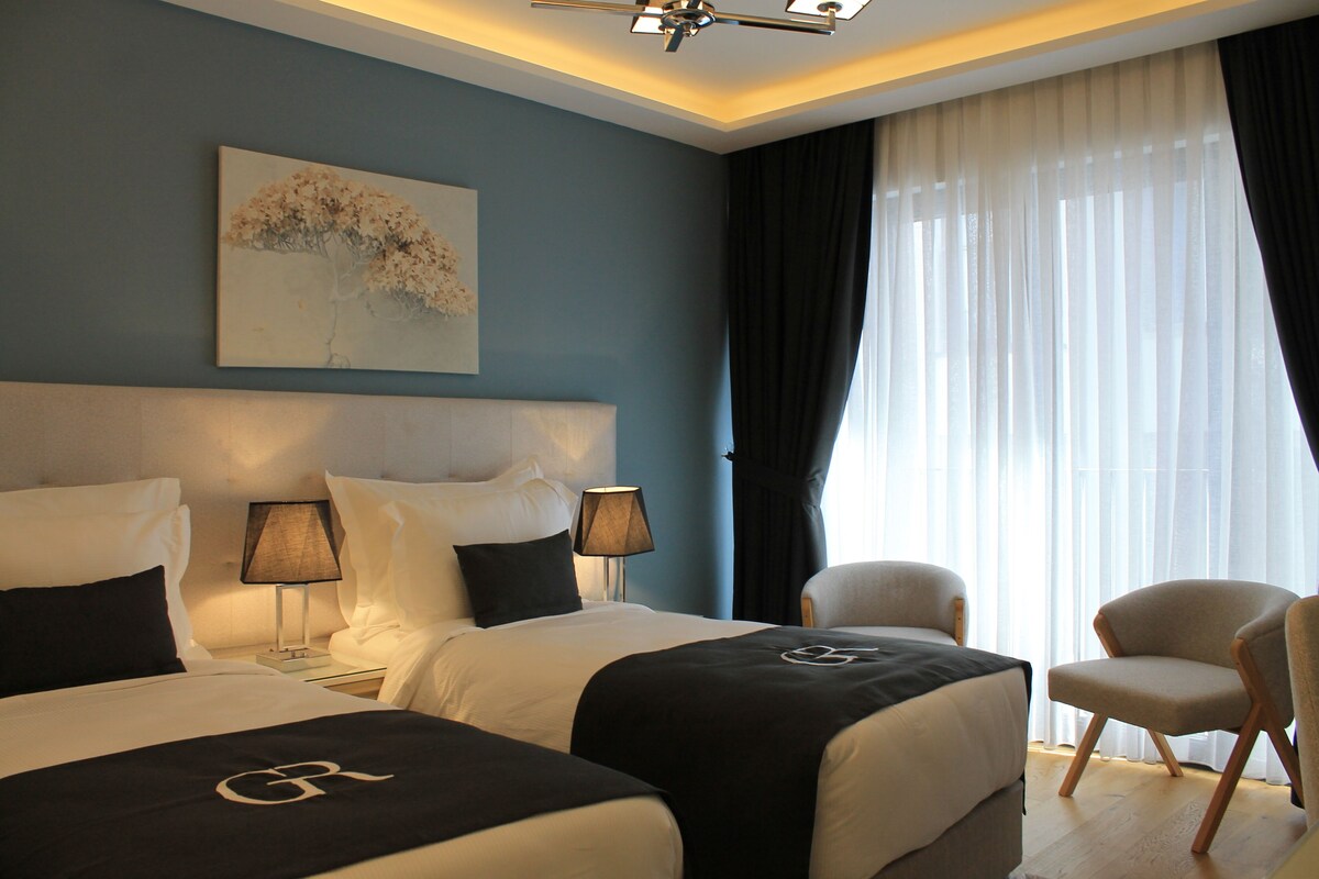 Une chambre d’hôtel avec deux lits, une tête de lit, une table de chevet avec une lampe, une chaise et une grande fenêtre avec des rideaux légers.