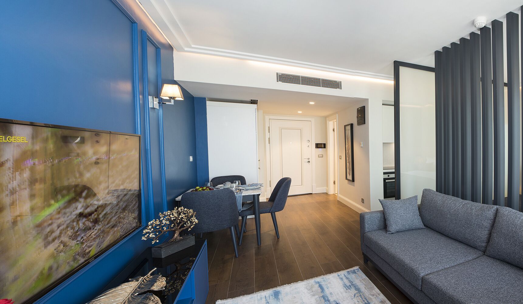 A modern living room with a large flat-screen TV, a grey sofa, and a dining area with a table and chairs.