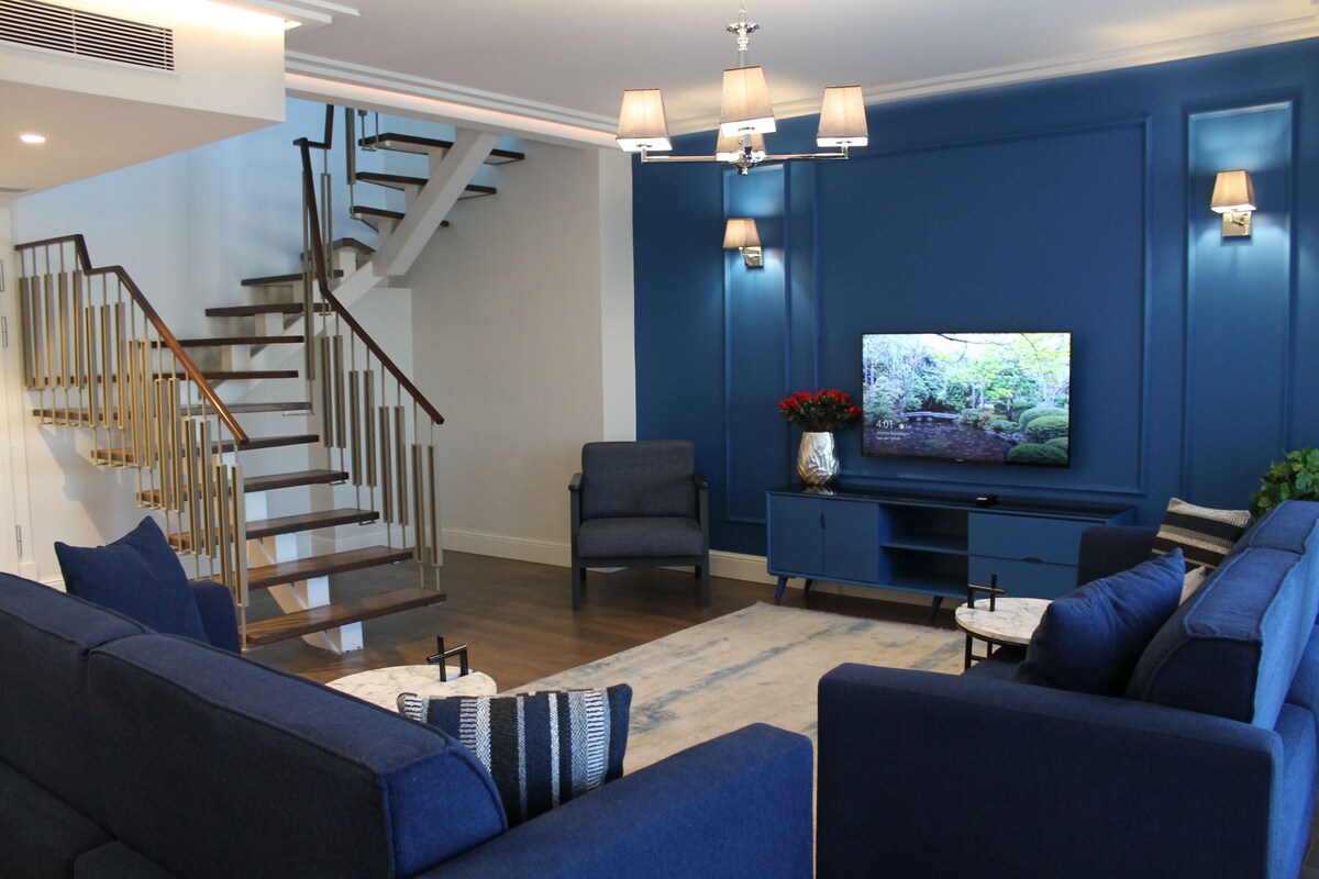 Un salon moderne avec un mur d’accent bleu, un téléviseur à écran plat, un escalier et un coin salon comprenant des canapés et une table basse.