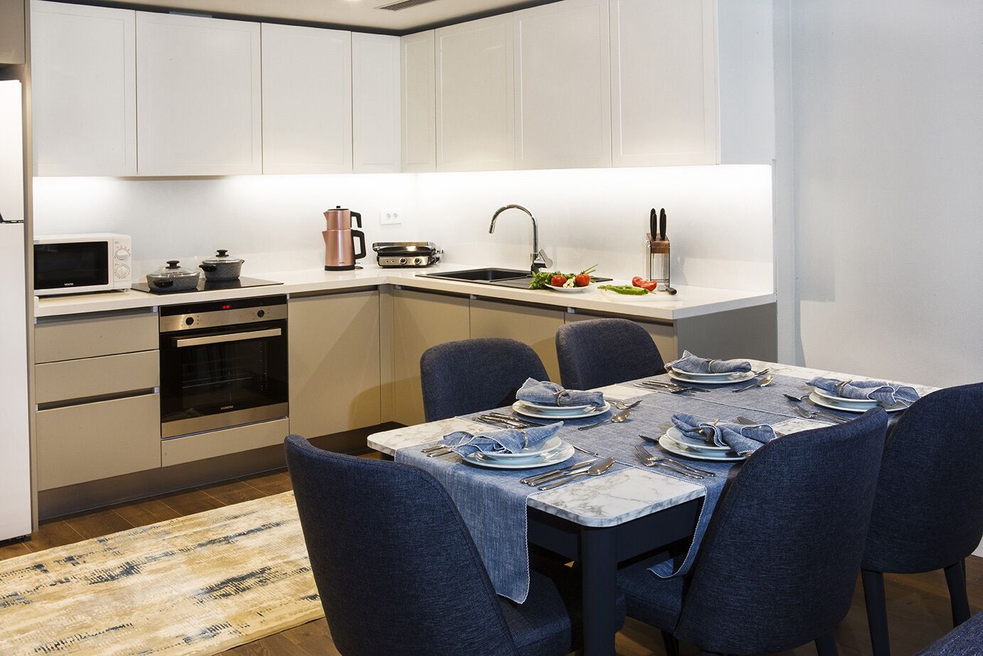Cocina moderna con armarios blancos, horno empotrado, microondas, hervidor y pileta. Se aprecia una mesa de comedor con platos, cubiertos y un camino de mesa.