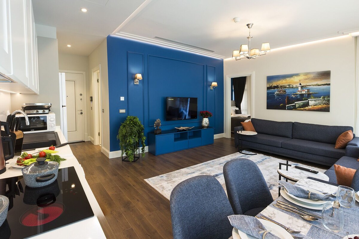 Un salon moderne avec un mur d’accent bleu, un téléviseur à écran plat, un canapé et un coin repas avec une table mise pour deux personnes.
