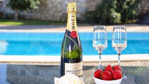 Een fles Moët & Chandon champagne, twee glazen en een schaal met aardbeien op een tafel bij een zwembad, met een stenen gebouw op de achtergrond.