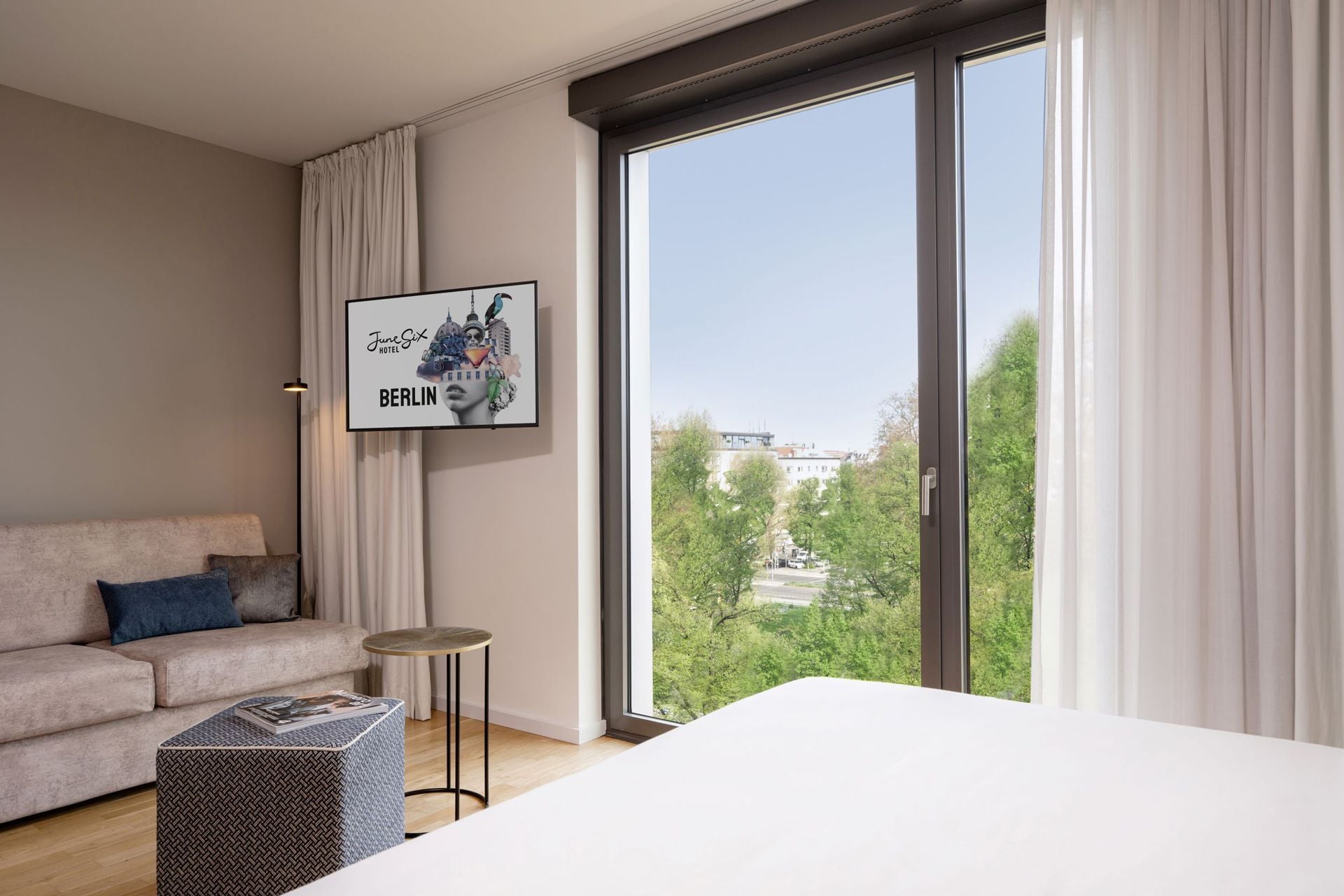 Ein modernes Hotelzimmer mit großem Fenster, einem gerahmten Bild von Berlin, einer Couch, einem kleinen runden Tisch und einem gemusterten Hocker.
