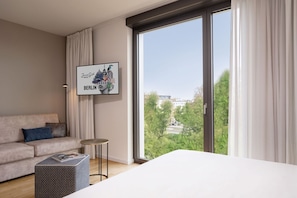 Una habitación de hotel moderna con un ventanal grande, un cuadro enmarcado de Berlín, un sofá, una mesita redonda y un otomano estampado.