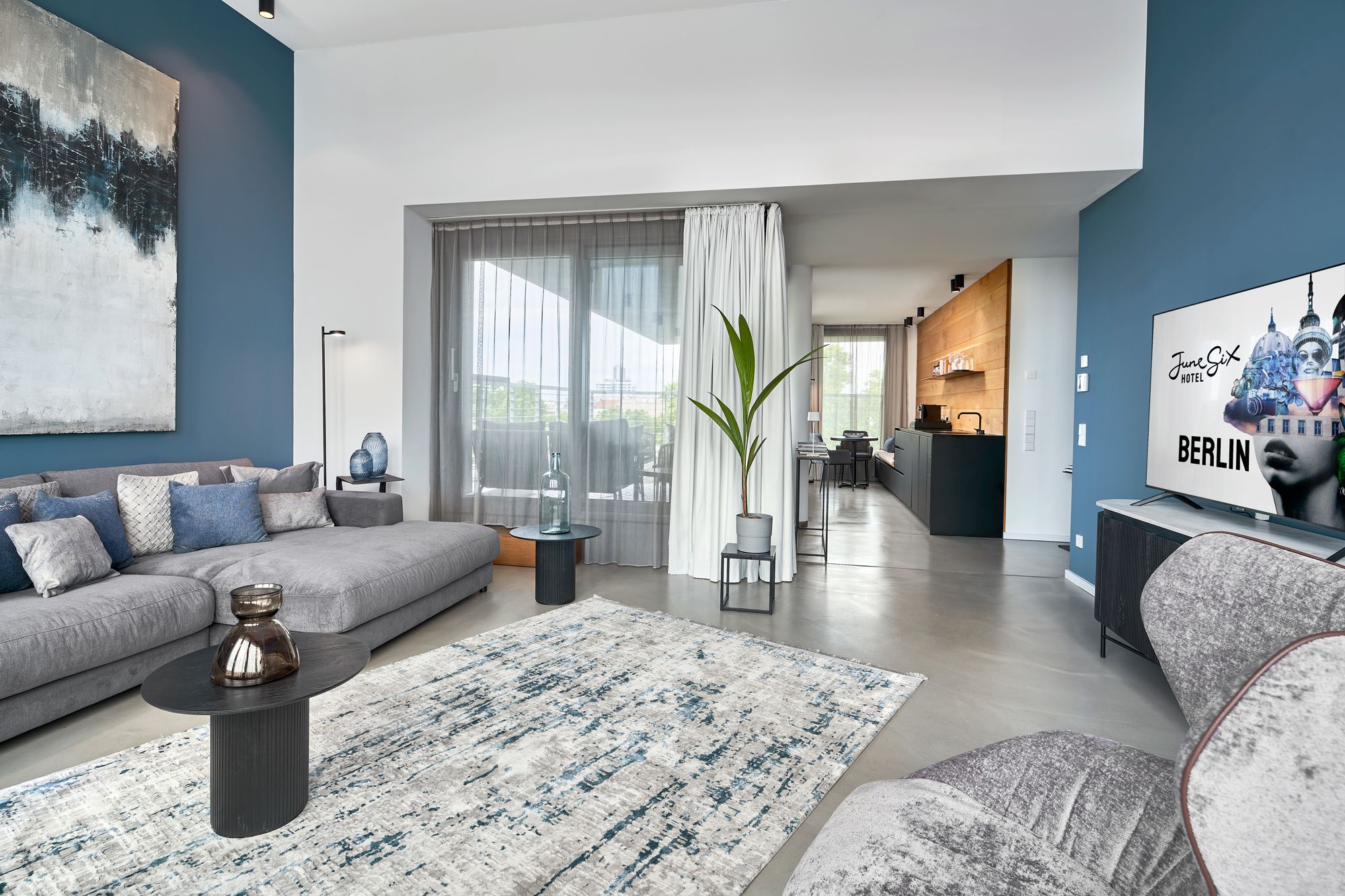 Un salon moderne avec un canapé gris, une table basse et un grand tableau. On aperçoit également un coin repas avec une cuisine en arrière-plan.