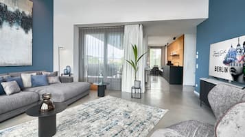 Un salon moderne avec un canapé gris, une table basse et un grand tableau. On aperçoit également un coin repas avec une cuisine en arrière-plan.