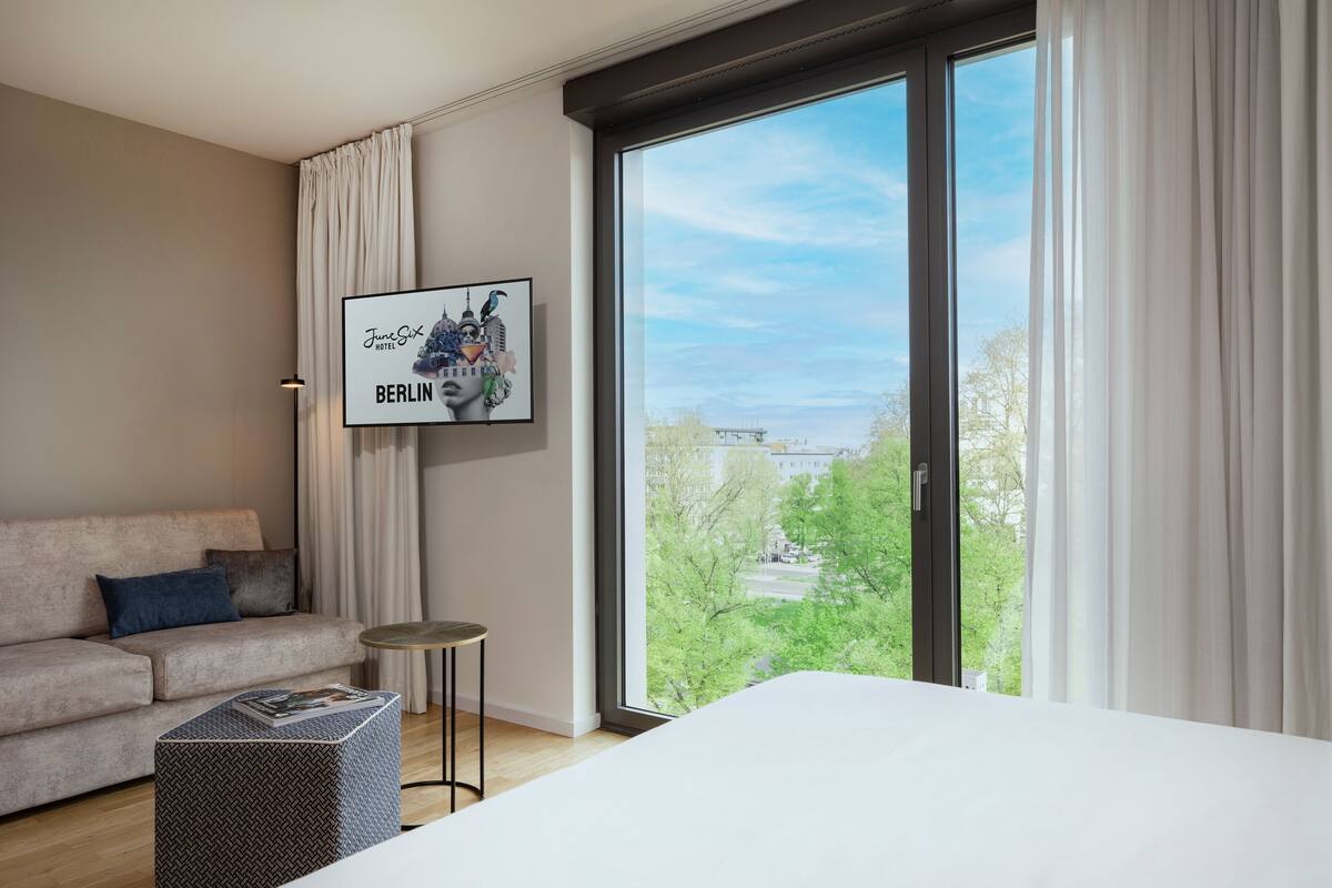 Une chambre d’hôtel moderne avec une grande fenêtre, un tableau encadré représentant Berlin, un canapé et une petite table d’appoint.