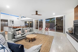 A modern living room with a grey sofa, a round coffee table, and two armchairs. There is a kitchen area with stainless steel appliances and a dining table. The room has large sliding glass doors leading to an outdoor area.