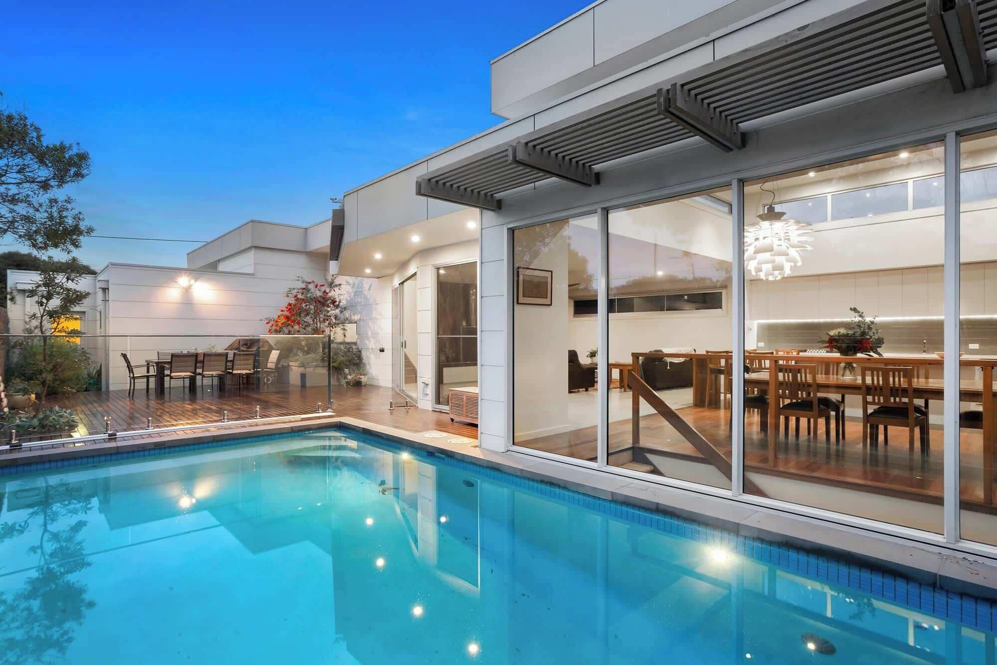 A modern house with a swimming pool, large glass doors leading to a dining area, and a well-lit outdoor space.