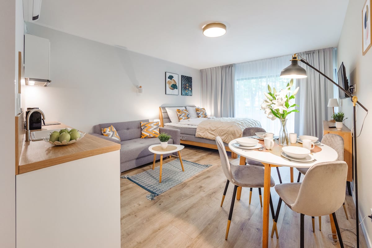 A modern, well-lit bedroom with a bed, bedside table, and a nightstand. There is a dining area with a table set for two, chairs, and a vase with flowers.