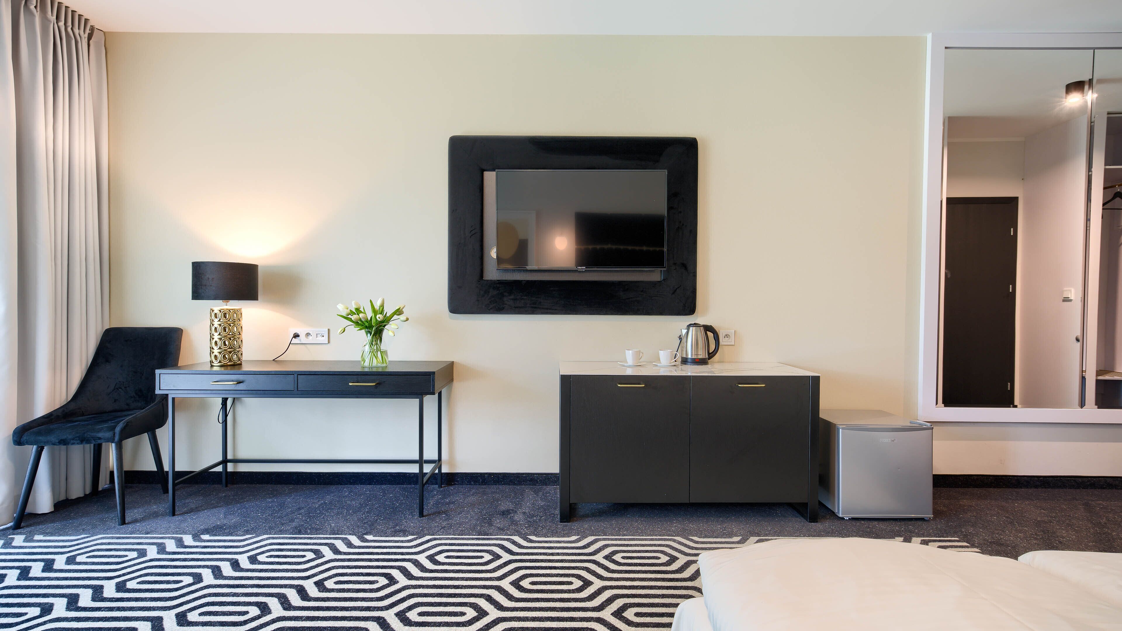 Ein Hotelzimmer mit Bett, Schreibtisch mit Lampe, Fernseher, Minibar und einem durch eine Glastür einsehbaren Badezimmer.