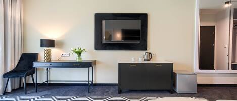Ein Hotelzimmer mit Bett, Schreibtisch mit Lampe, Fernseher, Minibar und einem durch eine Glastür einsehbaren Badezimmer.