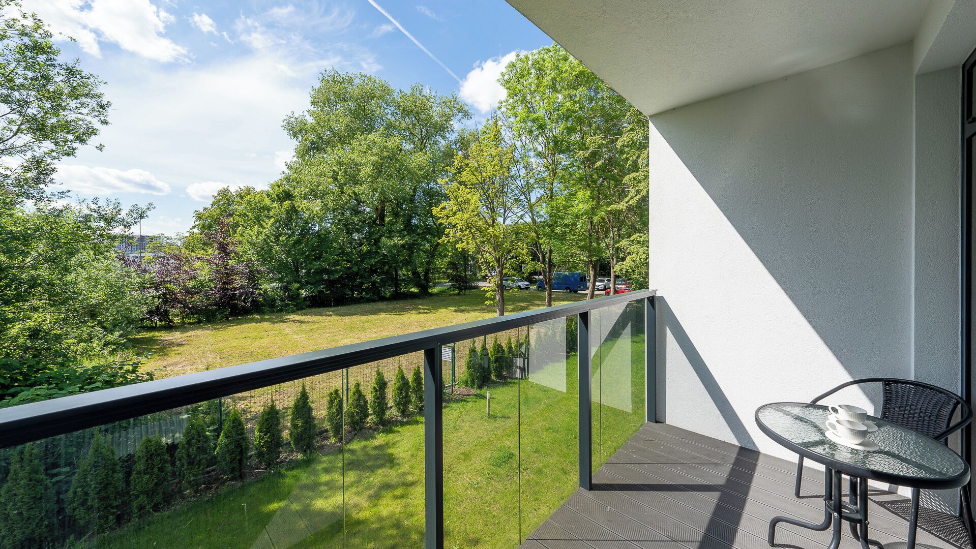 Ein Balkon mit Glasgeländer, der auf eine Rasenfläche mit Bäumen und geparkten Autos blickt.