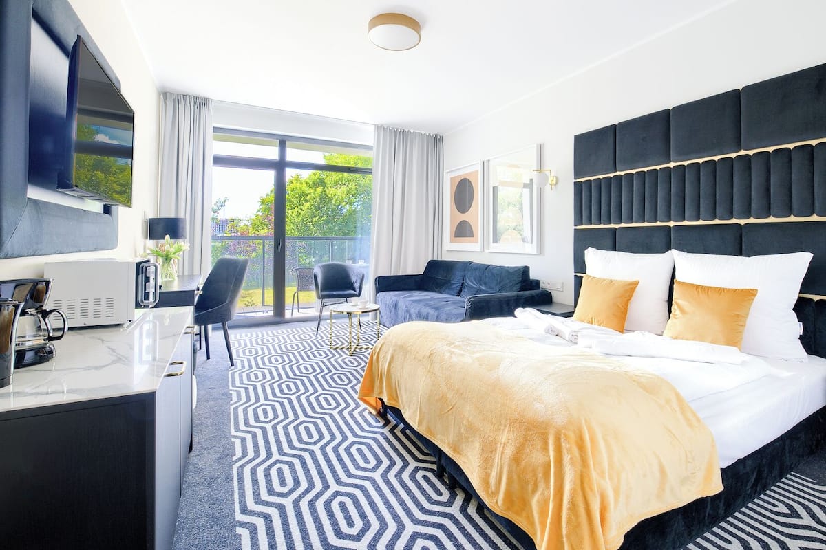 A modern hotel room with a large bed, a flat-screen TV, a desk with a coffee maker, and a balcony with a view of greenery.