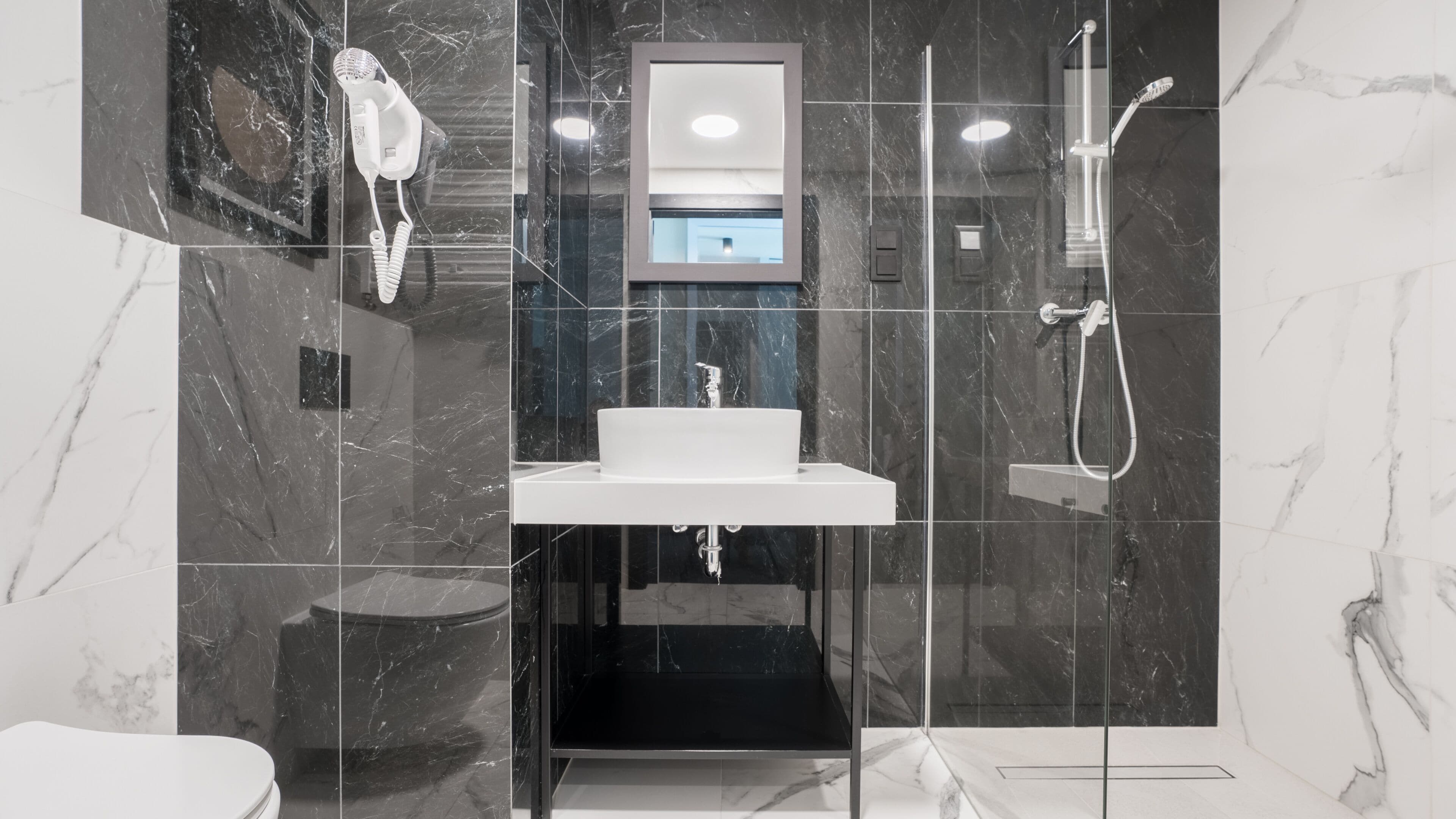Un baño moderno con suelo de mármol, lavabo blanco, ducha con mampara de vidrio y secador de pelo montado en la pared.