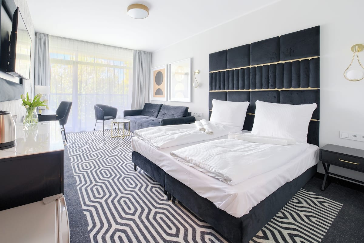 A modern hotel room with a large bed, a desk with a kettle, a sofa, and a patterned rug.