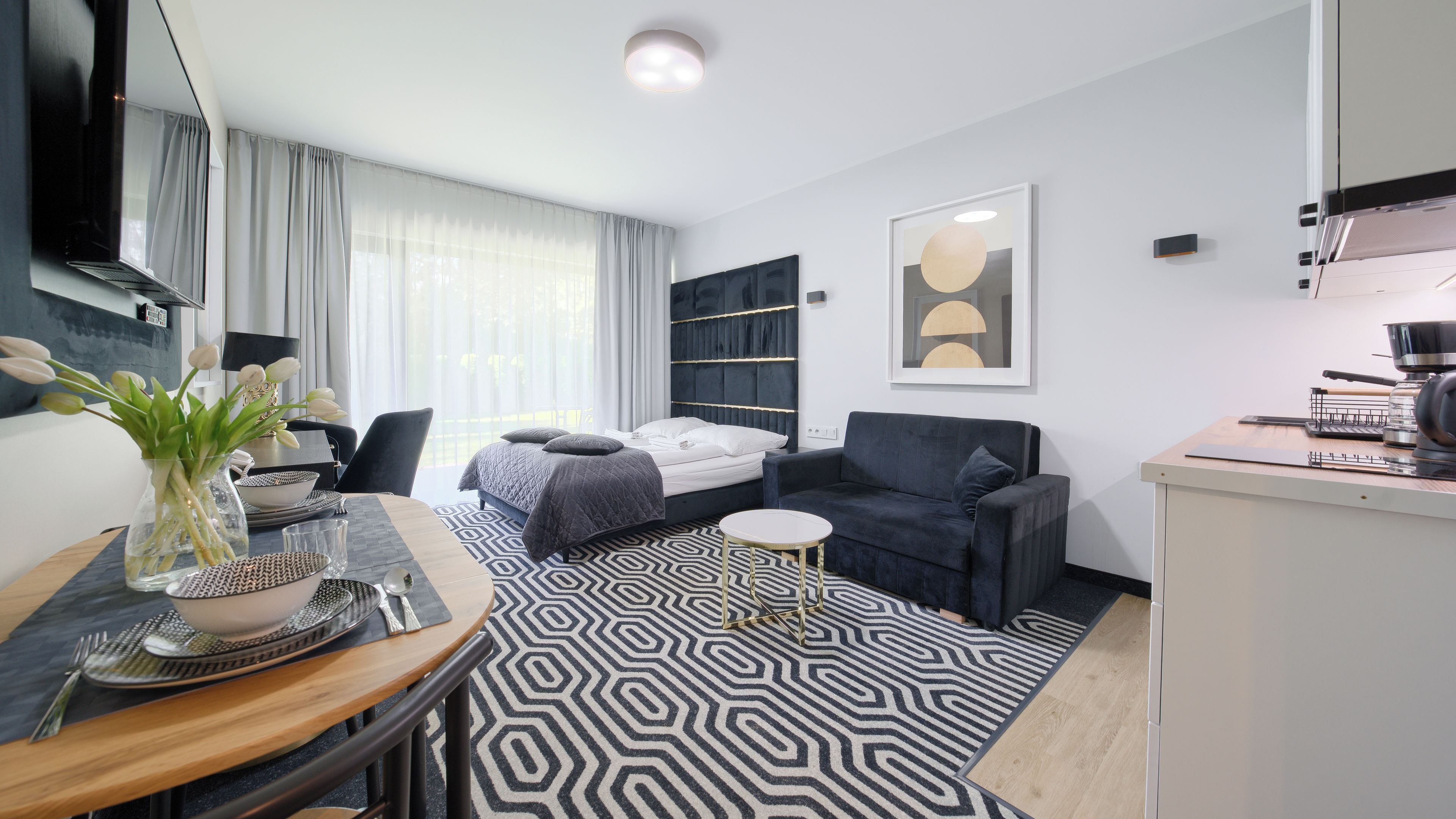 A modern hotel room with a bed, sofa, dining table, and kitchenette.