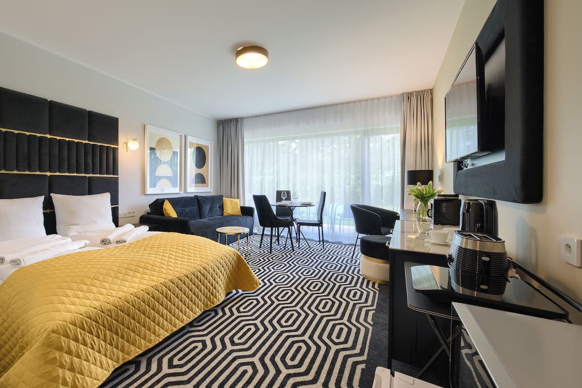 A hotel room with a large bed, a desk with a coffee maker, a TV, and a dining area with chairs and a table.
