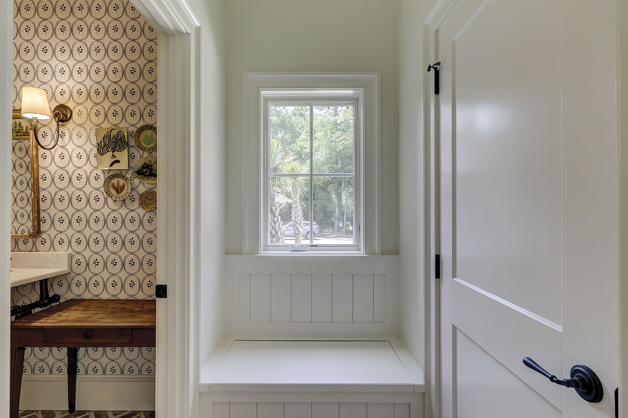 A bathroom with a white bench, a window with a view of trees, and patterned wallpaper.
