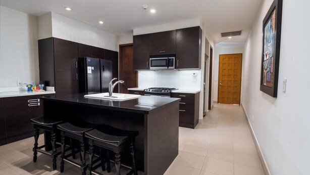 Una cocina moderna con armarios oscuros, una isla central con taburetes, un lavamanos de acero inoxidable y un microondas sobre la estufa.