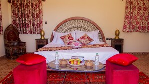 Una habitación tradicional con una cama grande, dos otomanas rojas, una bandeja con frutas y botellas de agua, y decoraciones de pared ornamentadas.