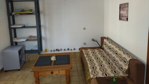 A small, furnished room with a wooden table, a sofa, a shelf with folded clothes, and a wall-mounted picture. - Charming studio 25 min from vallon pont d'arc (Le Garn)
