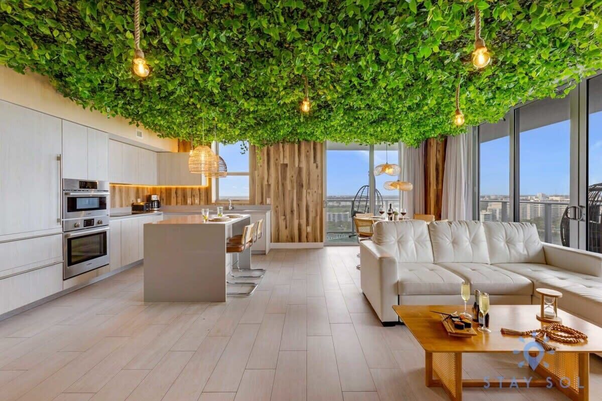 A modern kitchen with white cabinets and stainless steel appliances, a wooden dining area, and a living space with a white sofa and a coffee table.