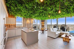 A modern living room with a green leafy ceiling, a kitchen area, and a view of the city.