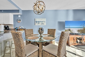 A dining area with a glass table surrounded by woven chairs, a decorative centerpiece, and a beach view through a large window.