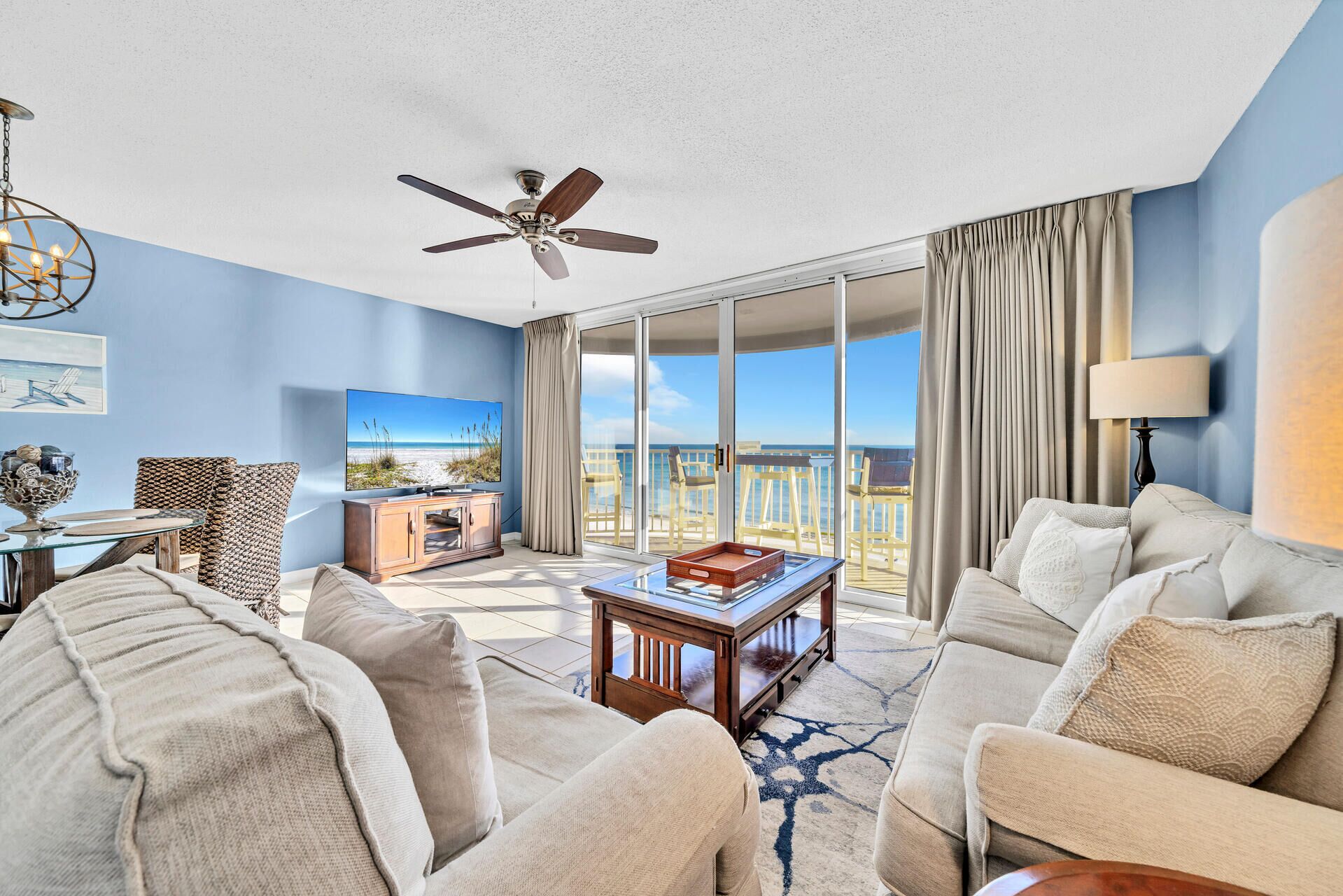 A spacious living room with a large TV, comfortable seating, a ceiling fan, and a view of the ocean through large sliding doors.