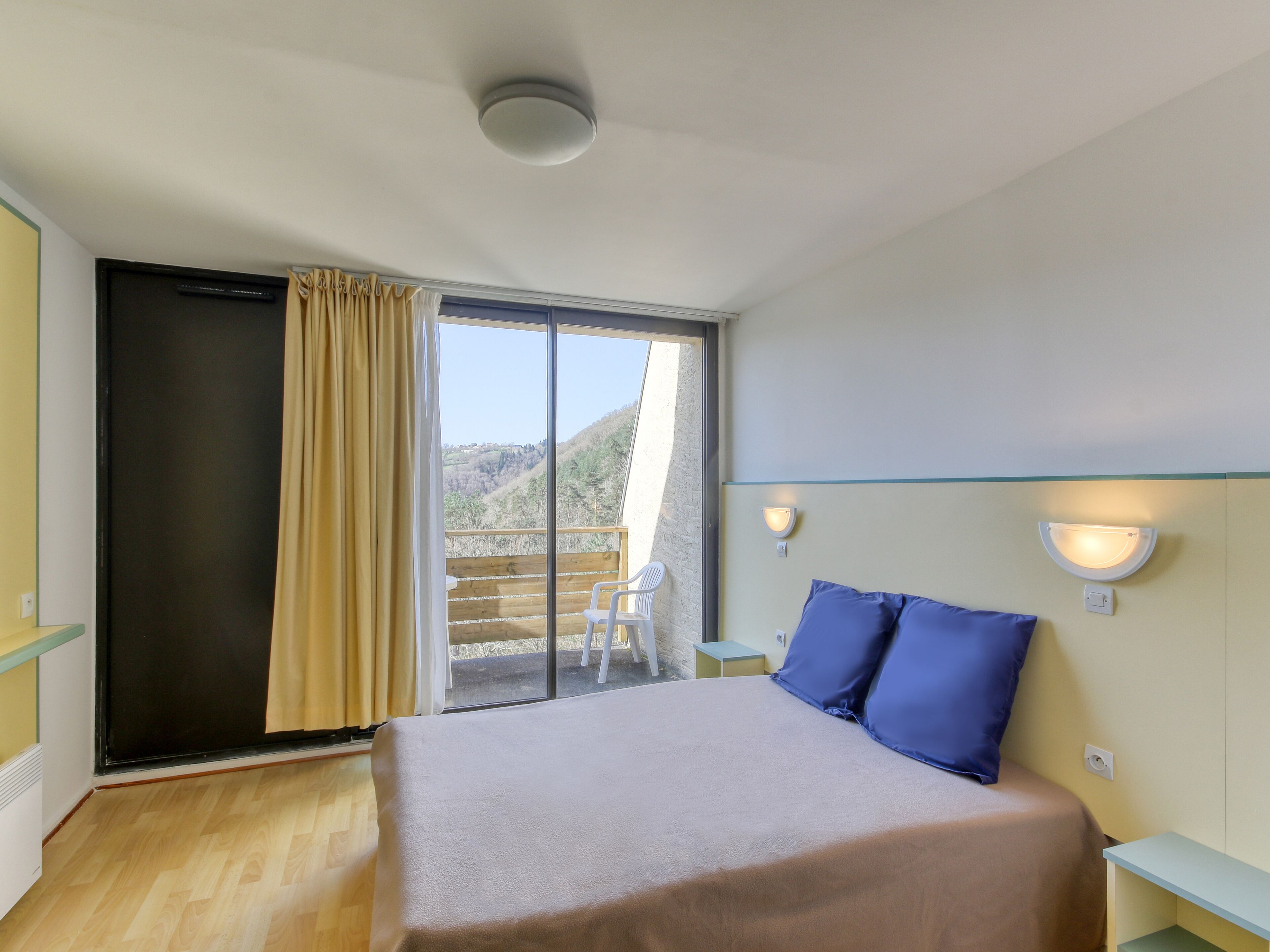 Un dormitorio con cama, ventana con cortinas, vista al exterior e iluminación en el techo.