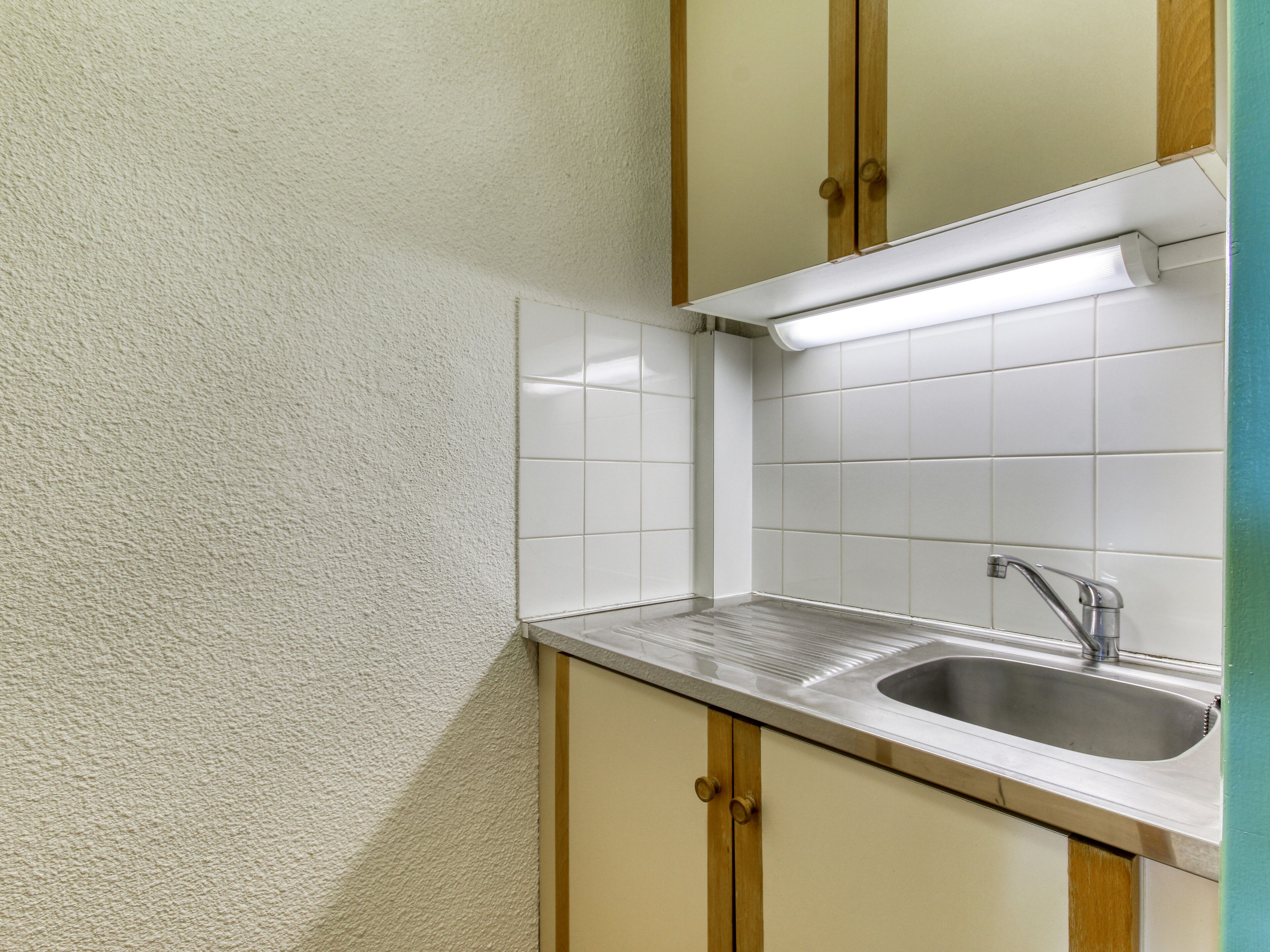 A small kitchen with white tiles, a stainless steel sink, and wooden cabinets.