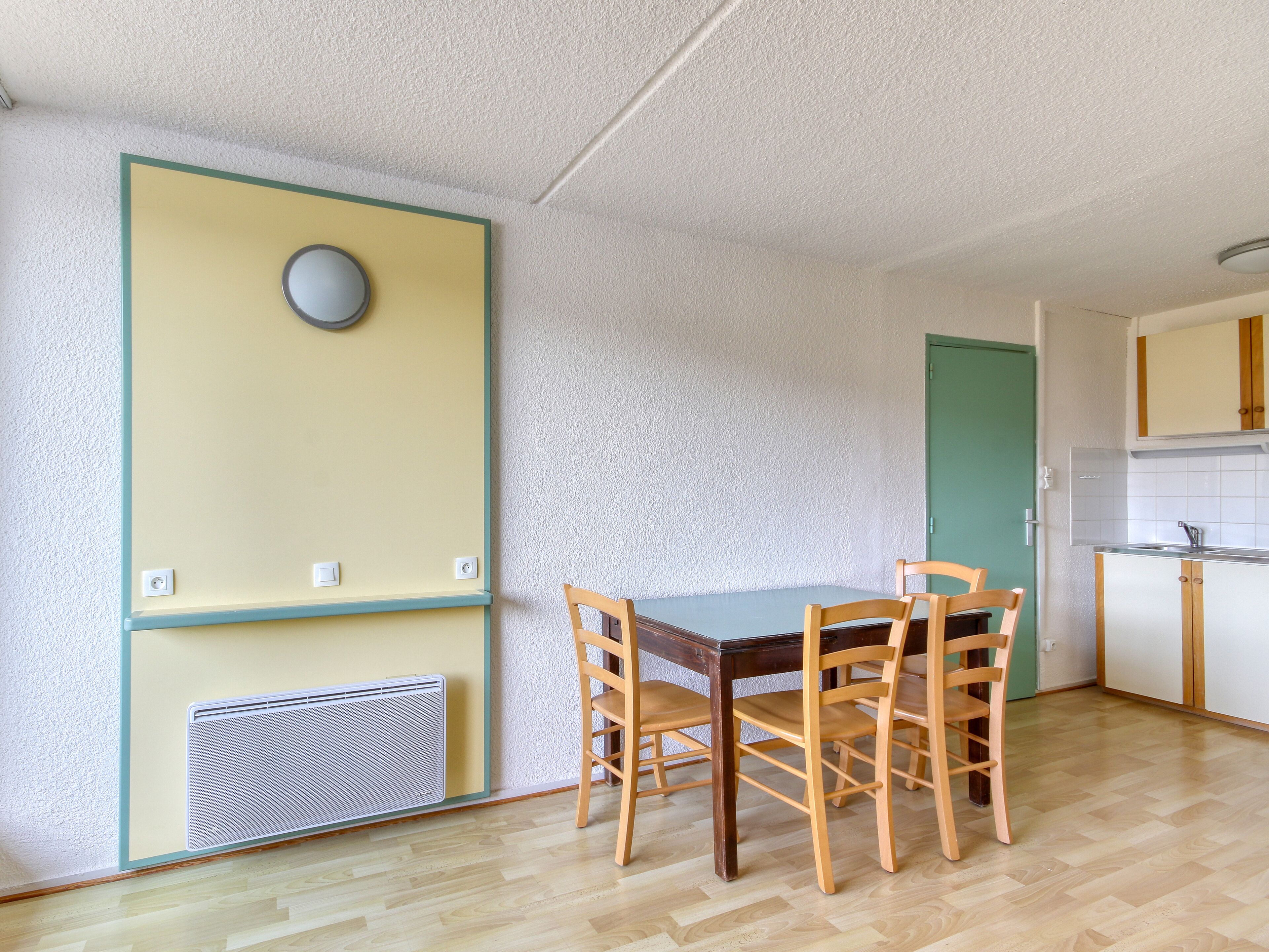 A small, well-lit room with a wooden dining table and chairs, a radiator, a green door, and a kitchenette with white cabinets.