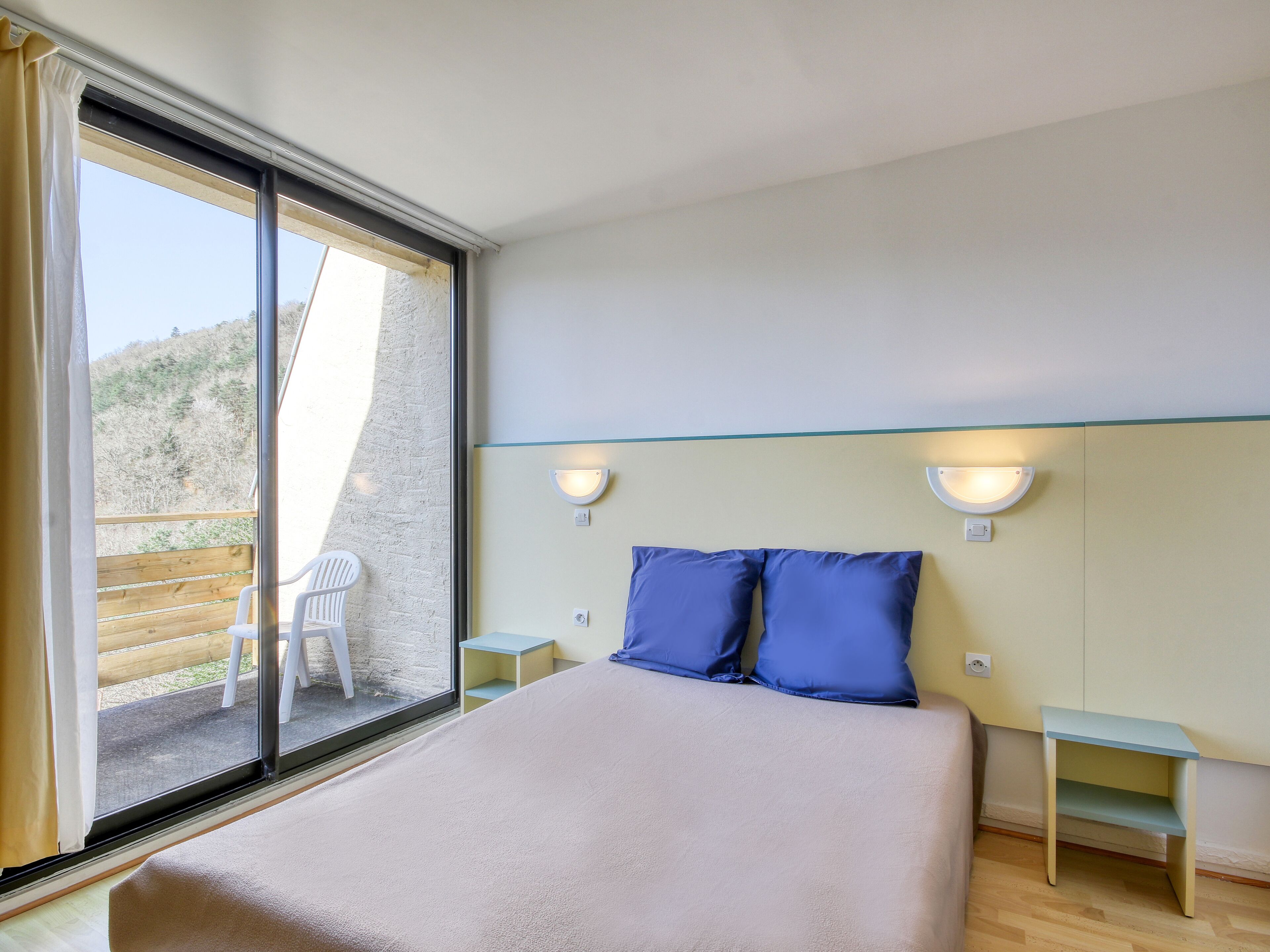 A bedroom with a large bed, two nightstands, a window with curtains, and a view of a wooden deck.