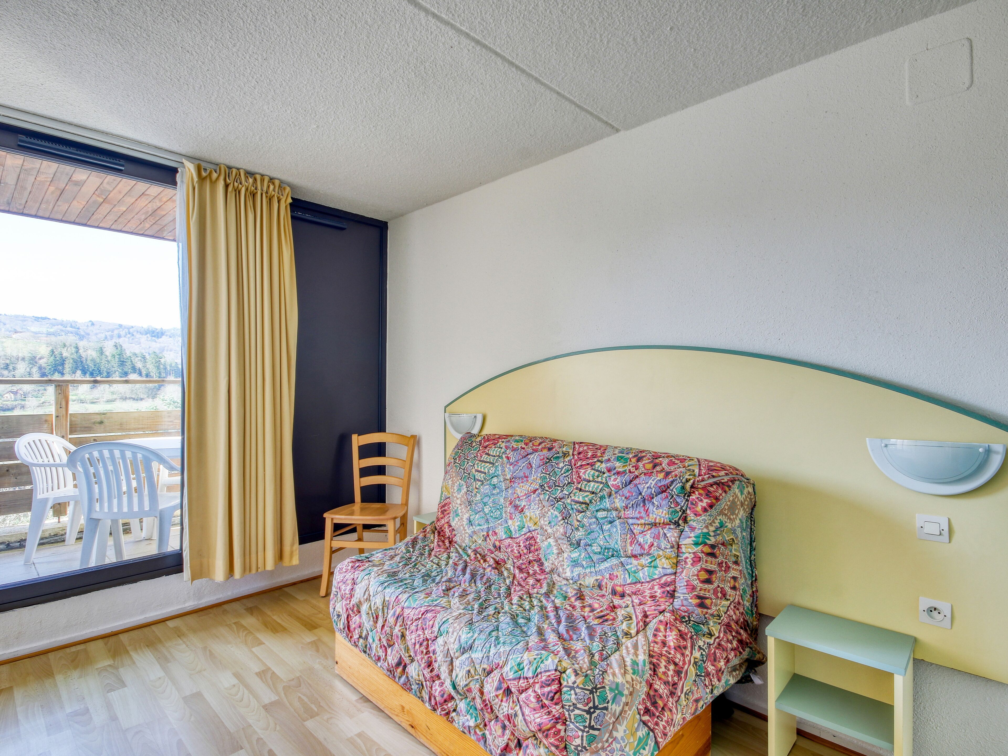 A bedroom with a bed, a chair, a small table, a window with curtains, and a view of the outdoors.