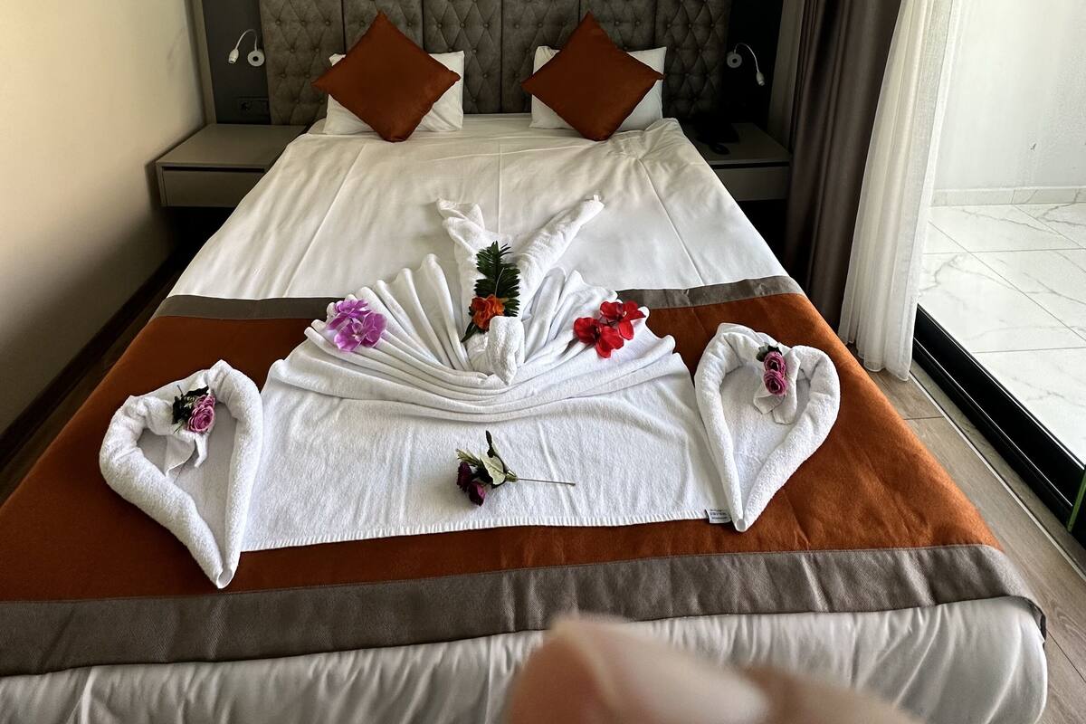Ein Hotelzimmer mit einem großen Bett, zwei Kissen, einem Kopfteil, einem Nachttisch und einem Fenster mit Vorhängen.