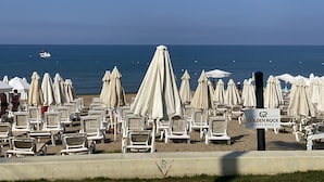 Blick auf einen Strand mit Reihen aus weißen Liegestühlen und Sonnenschirmen, einem Schild für das Side Golden Rock Hotel & Spa und einem Boot in der Ferne.